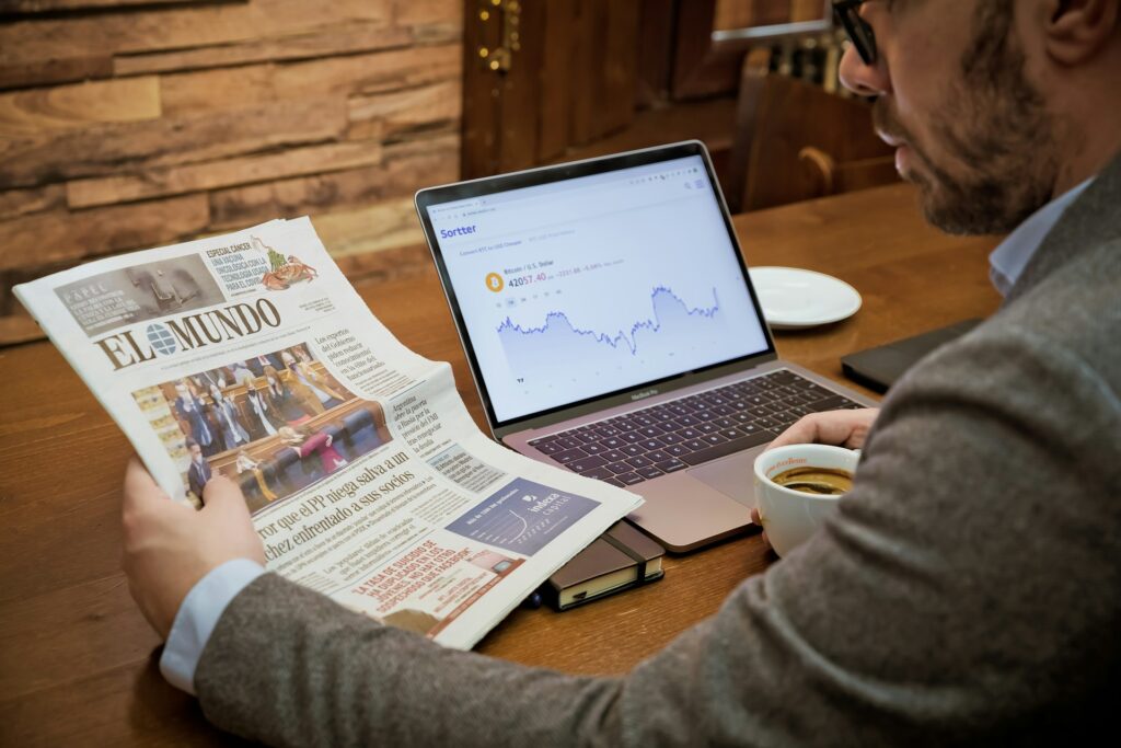 A man reading a newspaper from Spain with a computer opened showing a stock graph
