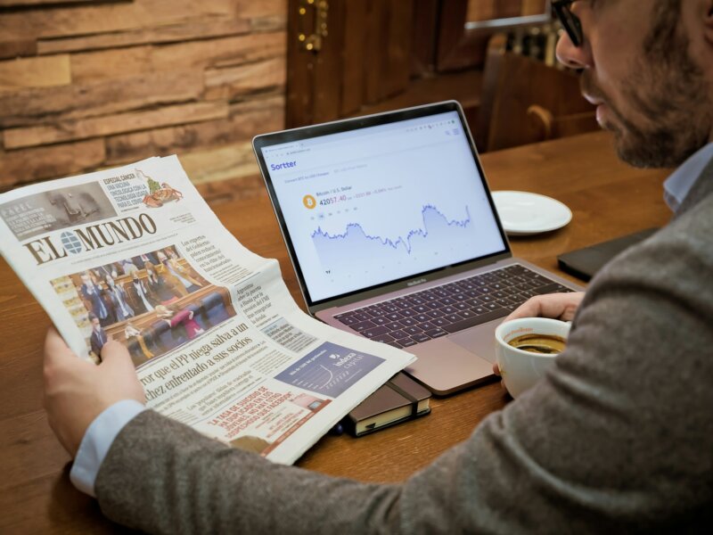 A man reading a newspaper from Spain with a computer opened showing a stock graph